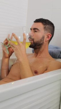 Yetişkin beyaz bir adam sarı bir içecek içerken küvette dinleniyor.