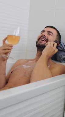 Yetişkin beyaz bir adam telefonda konuşurken küvette dinleniyor ve elinde bir bardak içki tutuyor.