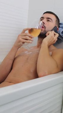 Yakışıklı adam bir bardak içki ve telefonla küvette dinleniyor.