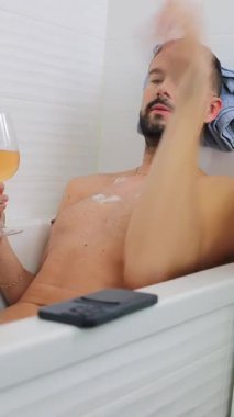 Genç adam bir bardak içki ve telefonla küvette dinleniyor.