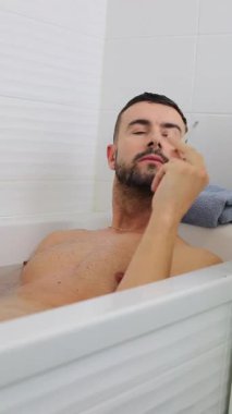 Genç adam, düşünceli bir ifade ve sigarayla küvette dinleniyor.