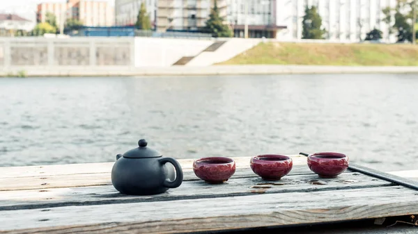 Black clay kettle and three burgundy ceramic saws on the waterfront in the city, the Chinese traditional tea drinking. Close-up image with blurred background.