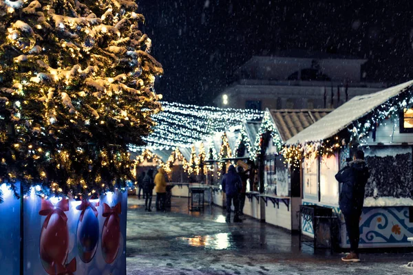 Fiesta de Navidad y Año Nuevo plaza de feria en la ciudad por la noche, nevada, árbol de Año Nuevo y casas con guirnaldas. — Foto de Stock