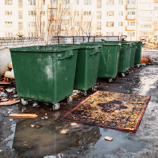 Bidoni della spazzatura in un cortile provinciale russo e un tappeto chic di fronte a loro sdraiato in una pozzanghera — Foto Stock