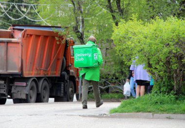 24.05.2020. Syktyvkar, Rusya. Yemek dağıtım şirketinin kuryesi 'Teslimat Kulübü' nün arkasında yeşil bir çanta ve yeşil üniformalarla ilkbaharda şehir caddesinde bir kamyon ve ağaçların önünde yürüyor..