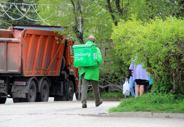 24.05.2020. Syktyvkar, Rusya. Yemek dağıtım şirketinin kuryesi 'Teslimat Kulübü' nün arkasında yeşil bir çanta ve yeşil üniformalarla ilkbaharda şehir caddesinde bir kamyon ve ağaçların önünde yürüyor..