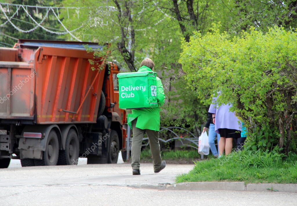 24.05.2020. Syktyvkar, Russia. The courier of the food delivery company 'Delivery club' with a green bag behind his back and in green uniforms walks down the city street in the spring against the backdrop of a truck and trees.