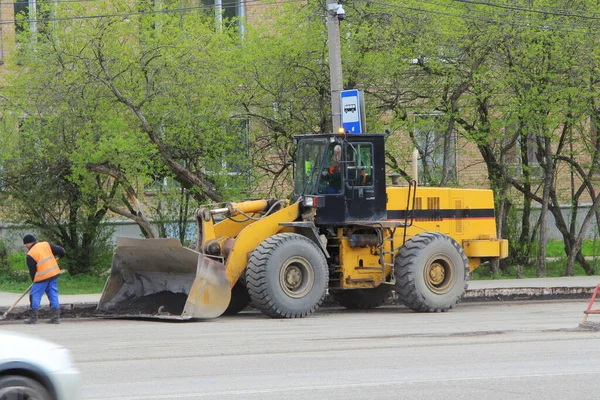 トラクターローダーは大きな黒い鉄のバケツとキャビンが付いている黄色です ロシアの街の通りに道路設備 春の道路補修工事 — ストック写真