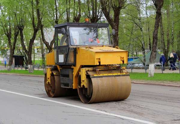 The asphalt rink is yellow with iron wheels on the road in the city against the background of green trees. Repair work on the street on laying asphalt.