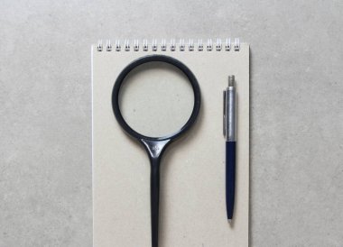 Üzerinde tükenmez kalem ve açık gri beton arka planda siyah büyüteç bulunan bir kağıt kapaklı bahar defteri. Yeni fikir, iş planı ve strateji kavramı, içeriğin geliştirilmesi ve uygulanması. Metin için boş boşluk.