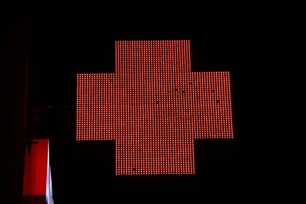 Glowing pixel cross of LED lamps on black background. Red cross symbol of health, medicine, self-care. Stock photo with empty space for text and design.