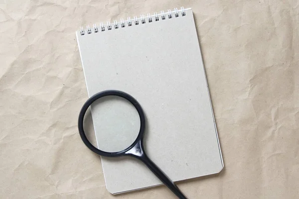 Gray notepad with white coiled spring and magnifier on a background of beige crumpled craft paper. With empty space for text and design