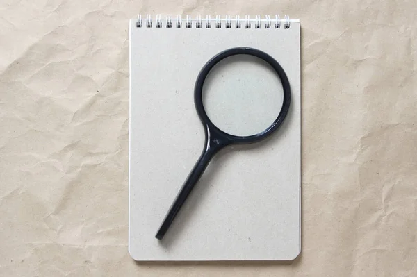 Gray notepad with white coiled spring and magnifier on a background of beige crumpled craft paper. With empty space for text and design