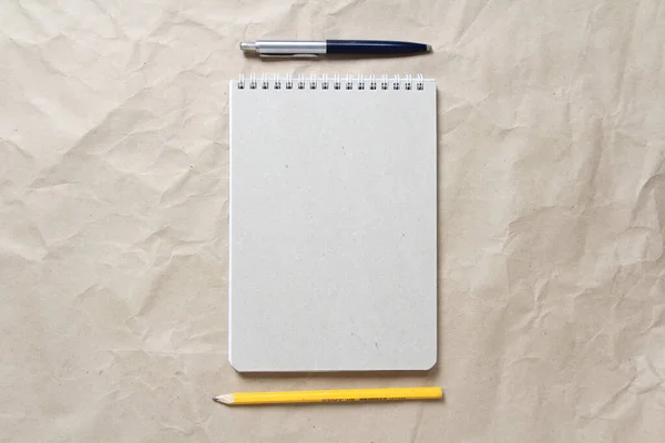 Gray notepad with white coiled spring and pen and pencil on a background of beige crumpled craft paper. With empty space for text and design
