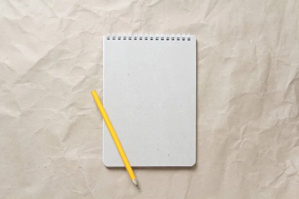 Gray notepad with white coiled spring and pencil on a background of beige crumpled craft paper. With empty space for text and design
