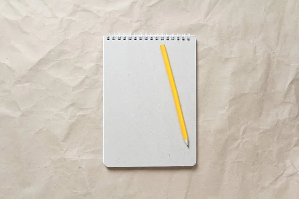 Gray notepad with white coiled spring and pencil on a background of beige crumpled craft paper. With empty space for text and design