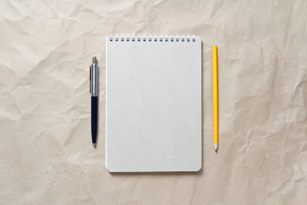 Γκρι σημειωματάριο με λευκό τυλιγμένο ελατήριο και στυλό και μολύβι σε φόντο από μπεζ τσαλακωμένο χαρτί χειροτεχνίας — Φωτογραφία Αρχείου