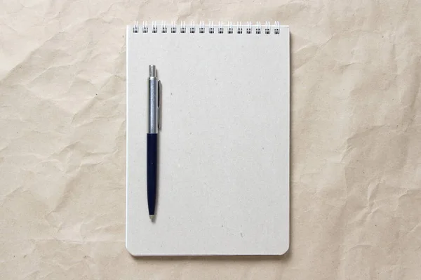 Gray notepad with white coiled spring and pen on a background of beige crumpled craft paper