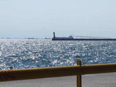 Sakin sular parlak güneş ışığı altında deniz fenerinin bulunduğu uzun bir rıhtımın yanında parlar. Gemiler uzaktan görülebilir, sakin atmosferi güçlendirir..