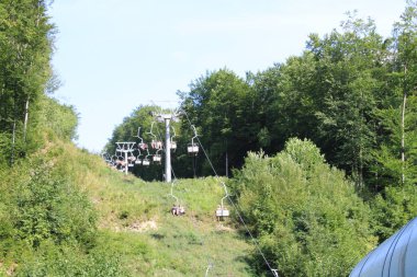 Boş bir kayak merkezi, sık ormanlarla çevrili yemyeşil bir yamaçtan yükseliyor. Parlak güneş, yaz yapraklarının canlı renklerini güçlendirir..