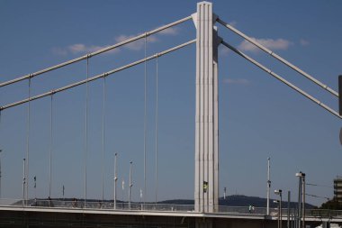 Kablo destekli bir köprünün yüksek yapısı, modern tasarımını sergileyen canlı mavi gökyüzüne karşı belirgin bir şekilde sergilenmektedir. Güneş ışığı köprünün temiz hatlarını vurguluyor.