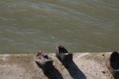 Bir çift yıpranmış ayakkabı, kısmen bulanık suya batmış bir nehir kıyısında duruyor. Güneş ışığı nehirden yansıyarak terk edilme havasını arttırır..