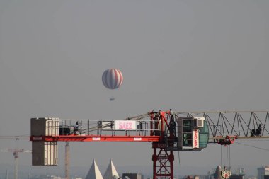 Canlı bir sıcak hava balonu inşaat vincinin üzerinde huzur içinde süzülür ve açık mavi gökyüzüne karşı çarpıcı bir kontrast yaratır. Şehir manzarası manzaraya derinlik katıyor..