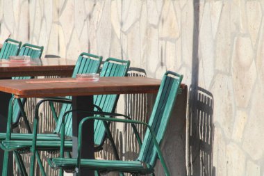 Konforlu bir açık hava kafe, güneşli taş bir duvara karşı düzgünce dizilmiş yeşil metal sandalyeler ve ahşap masalar içerir ve müşteriler için hoş geldin atmosferi yaratır..