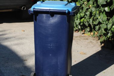 Mavi bir çöp kutusu beton bir yüzeyde duruyor, güneş ışığı gölgeleri yansıtıyor. Yemyeşil yeşillikler çöpün etrafını sarıp, canlı bir açık hava ortamı oluşturuyor..