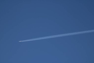 Bir jet uçağı, parlak gün ışığı altında el değmemiş mavi gökyüzünde uzun bir buhar izi bırakarak yükseklere uçar. Sakin atmosfer uçmak için açık bir gün olduğunu gösteriyor..