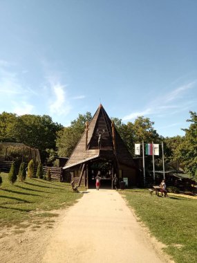 Bir grup ziyaretçi ağaçlarla çevrili büyük ahşap bir binaya doğru yol alıyorlar. Parkta güneşli bir yaz gününün tadını çıkarıyorlar..