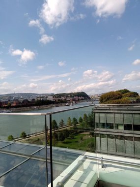 Berrak mavi bir gökyüzü, verimli yeşillik ve güneşli bir günde rıhtımda sıralanan modern binalarla şehri saran nehri vurgular..