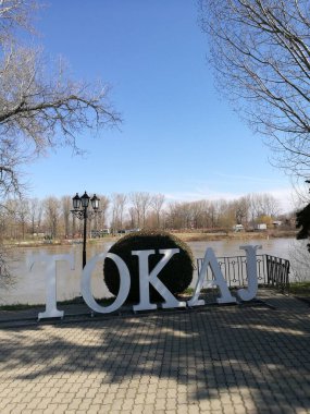 Tokaj tabelası, verimli yeşillikler ve ağaçlarla çevrili nehir kıyısında belirgin bir şekilde duruyor. Güneş tepemizde parlak bir şekilde parlarken, sakin bir atmosfer yaratıyor..