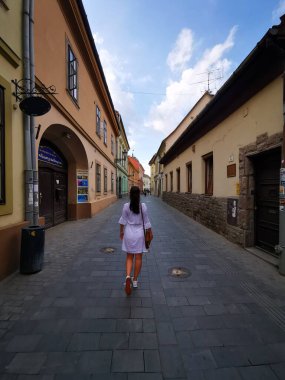 Beyaz elbiseli bir kadın, renkli binaların parlak mavi gökyüzünün altında sıralandığı eski bir kasabada, kaldırım taşı patika boyunca yürüyor..