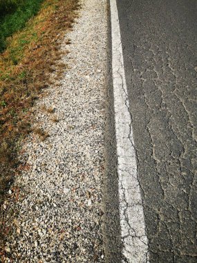 Yolun bir tarafında kurumuş çimenler, diğer tarafında da kırık asfalt var. Kırsal kesimi ve yıpranmış koşulları vurguluyor..