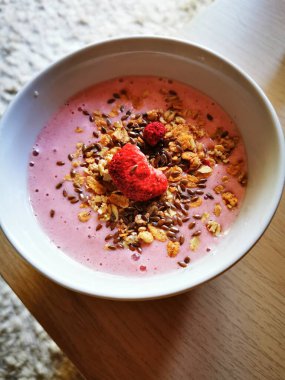 Pembe smoothie ile dolu renkli bir kase gevrek granola, keten tohumu ve taze ahududu içerir. Bu sağlıklı kahvaltı ahşap bir masada sergileniyor..