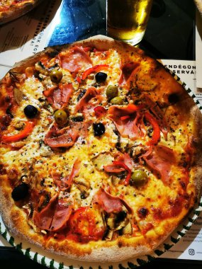 Taze pişmiş bir pizza, jambon, zeytin ve biber gibi çeşitli malzemelerle sunulmaktadır. Yakınlarda, bir bardak sarı içecek davetkar atmosfere katkı sağlar..