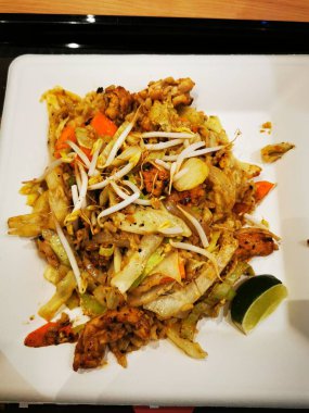 Tavuklu, sebzeli ve otlu kızarmış erişte servisi, limon kamaları eşliğinde, günlük yemek ortamında beyaz bir tabakta sunuluyor..