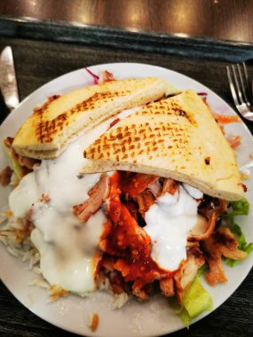 Yumuşak tavuk dilimleri kokulu pirinç ve taze salata üzerinde dinleniyor, yoğurt ve baharatlı sos serpiştirilmiş, sıcak ekmekle vurgulanmış, sıradan bir yemek atmosferinde..