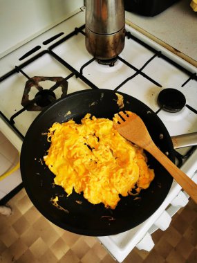 Lezzetli çırpılmış yumurtalar gaz sobasında siyah bir tavada hazırlanıyor. Sıcak ve davetkar bir mutfak ortamında, tavanın yanında duran ahşap bir spatula..