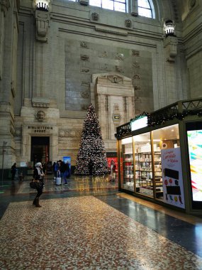 Güzel süslenmiş bir Noel ağacı, tarihi bir tren istasyonunda ayakta duruyor. Etrafı gezginlerle çevrili, şenlikli dekorasyonlar ve dükkanlar etrafında, sıcak bir tatil ortamı yaratıyor..