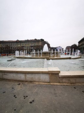Merkezi bir fıskiyeden gelen su jetleri, gri gökyüzünün altında tarihi mimariyle çevrili bir meydanda büyüleyici bir görüntü oluşturur..
