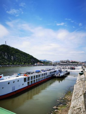 Açık bir günde sakin bir rıhtım boyunca rıhtıma yanaşan nehir tekneleri, bir tarafında yemyeşil bir yamaç, diğer tarafında parlak bir şehir silüeti bulunur..