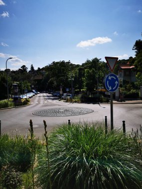 Parlak güneş ışığı, çimen ve ağaçlarla kaplı sakin bir kavşağı aydınlatıyor. Park halindeki arabalarla sakin bir ortam yaratıyor..