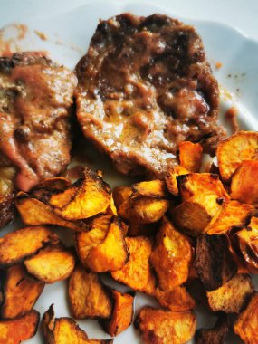 İçten bir yemek, yanında pişmiş et ve yanında da kızarmış patates dilimleri. Tabak basit beyaz bir arka planda, davetkar ve iştah açıcı..