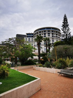 Bulutlu bir günde tarihi bir binanın yanında çeşitli tropikal bitkiler ve palmiyelerle dolu huzurlu bir bahçe yer alır ve sakin bir atmosfer yaratır..