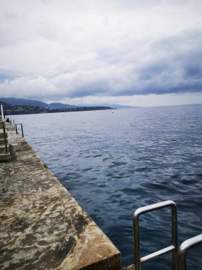 Sakin bir su kütlesi bulutların altında uzak bir kıyı şeridine doğru uzanıyor. Asfaltlı bir iskele, manzaranın tadını çıkarmak için huzurlu bir yer sunuyor..