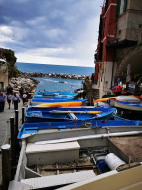 Parlak renkli tekneler kayalık kıyılarla çevrili bir limanda dinlenirken, turistler bulutlu bir gökyüzünün altında rıhtımda geziniyorlar..