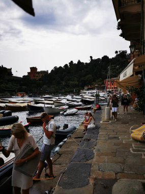 Portofino 'daki rıhtım çok sayıda tekneyle dolu ve turistler bulutlu bir gökyüzü altında taş kaldırım boyunca gezinerek canlı bir kıyı manzarası yaratıyorlar..