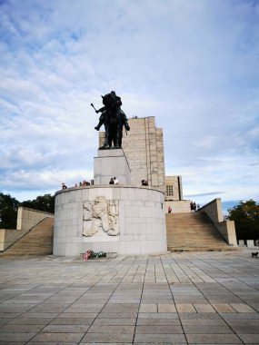 Büyük anıtta, ziyaretçilerle dolu geniş bir plazayla çevrili at sırtında dikilmiş tarihi bir figürün heykeli yer alıyor. Bulutlu gökyüzü dramatik bir zemin ekler.